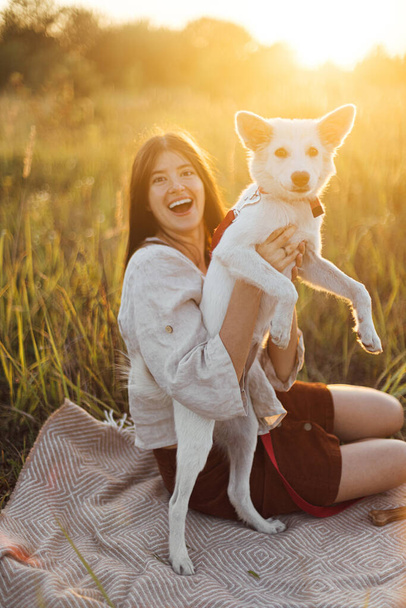 Stylish woman playing with her white dog on blanket in warm sunny light in summer meadow. Summer vacation and picnic with pet. Young boho female having fun with swiss shepherd puppy in sunset - Foto, afbeelding