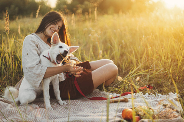 Stylish happy woman playing with her white dog with herb on blanket in warm sunny light in summer meadow. Summer vacation and picnic with pet. Young boho female relaxing with swiss shepherd puppy - Photo, Image