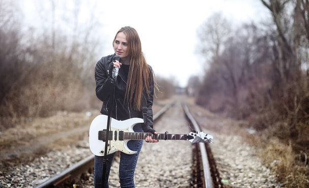 Beautiful young girl rocker with electric guitar. A rock musician girl in a leather jacket with a guitar sings. A rock band soloist plays the guitar and screams into microphone - Photo, Image
