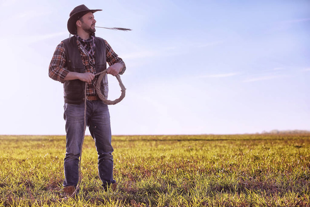 Kovboj v klobouku a laso stojící na poli při západu slunce - Fotografie, Obrázek