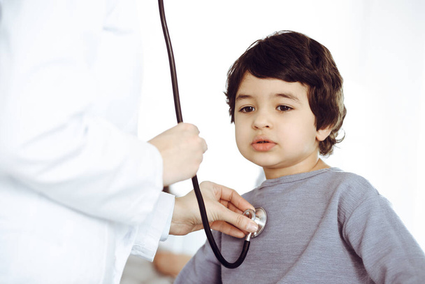 Doctor-woman examining a child patient by stethoscope. Cute arab boy and his brother at physician appointment. Medicine concept - Zdjęcie, obraz