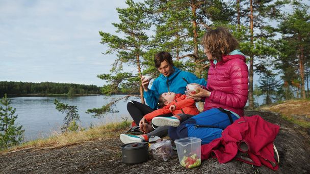4K Happy family of two young parents and pretty playful girl on spring camping vacation by lake near pine forest background eating breakfast from reusable tableware - Foto, afbeelding