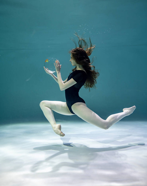 beautiful girl in a black swimsuit plays in orange fish underwater on a blue background - Foto, imagen