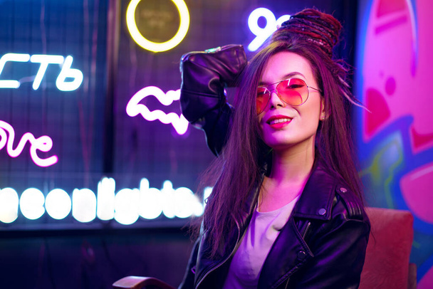 Fashion portrait of a young woman in sunglasses posing near neon signs in night club - Zdjęcie, obraz