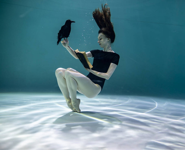  beautiful girl in a black swimsuit is enthusiastically reading a book underwater on a blue background - Фото, изображение