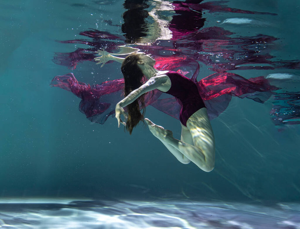   gyönyörű lány egy burgundi fürdőruhában pózol a víz alatt egy piros pareo a kék háttér - Fotó, kép