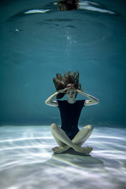 красивая девушка в черном трико делает гимнастические упражнения под водой на синем фоне - Фото, изображение
