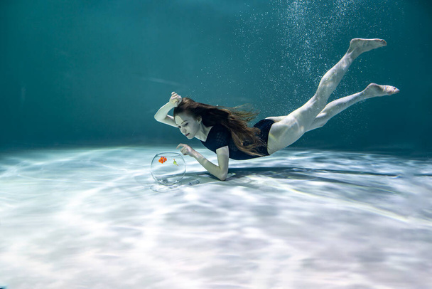 une belle fille en maillot de bain noir sous l'eau tient un aquarium avec des poissons dans ses mains et les admire - Photo, image