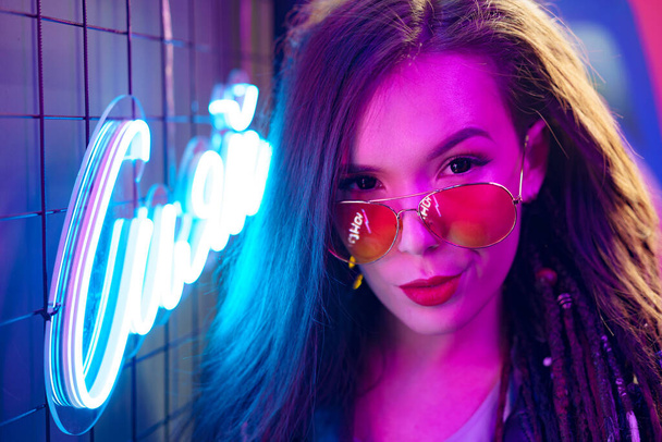 Fashion portrait of a young woman in sunglasses posing near neon signs in night club - Fotó, kép