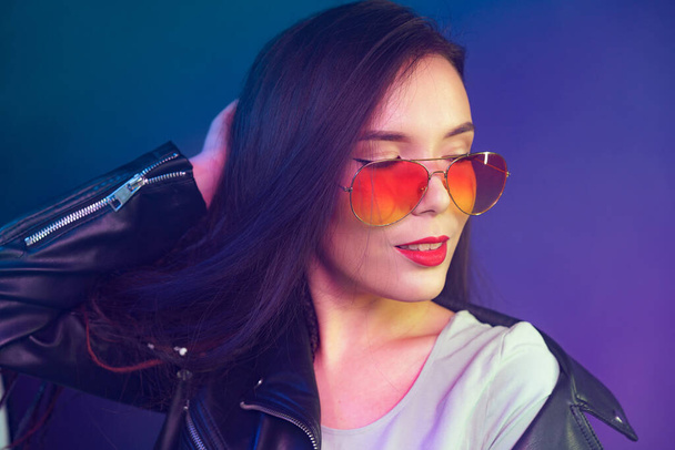 Asian girl in sunglasses and black leather jacket posing against dark blue background - Фото, изображение