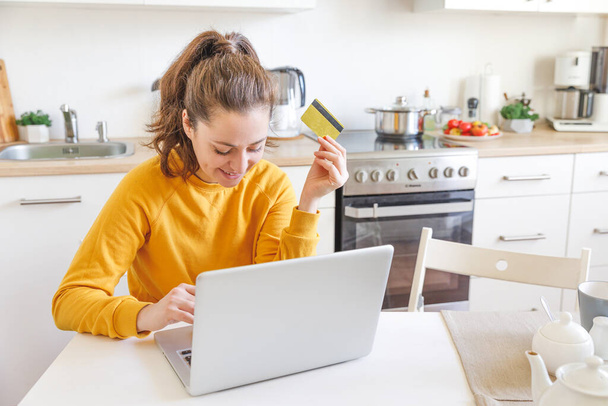 Kobieta robi zakupy online i płaci złotą kartą kredytową. Młoda dziewczyna siedzi z laptopem kupując w Internecie wprowadź dane karty kredytowej na tle kuchni wewnętrznej. Koncepcja e-commerce zakupów online - Zdjęcie, obraz