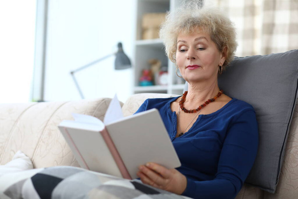 Elderly woman sitting on sofa and reading book at home - Photo, Image