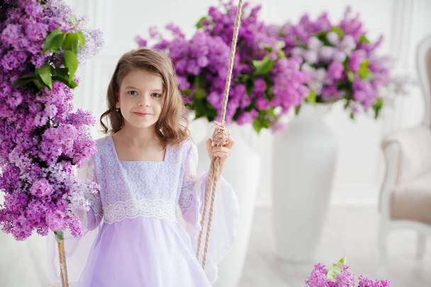 Hermosa niña de edad preescolar en un vestido delicado sentado en un columpio decorado con flores de color lila. - Foto, imagen