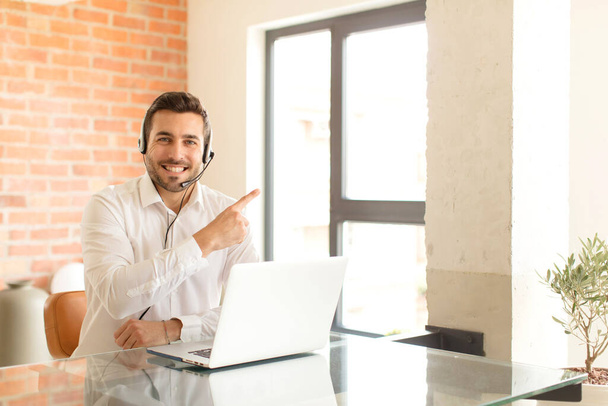 handsome telemarketer smiling cheerfully, feeling happy and pointing to the side and upwards, showing object in copy space - 写真・画像