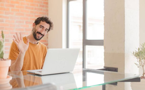 young bearded man smiling happily and cheerfully, waving hand, welcoming and greeting you, or saying goodbye and working with a laptop - Fotó, kép