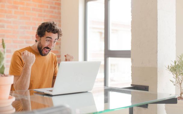 young bearded man shouting aggressively with an angry expression or with fists clenched celebrating success and working with a laptop - Foto, immagini