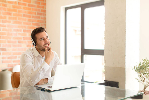 handsome telemarketer smiling with a happy, confident expression with hand on chin, wondering and looking to the side - Photo, image