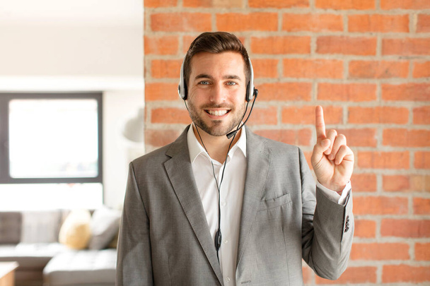 handsome telemarketer smiling and looking friendly, showing number one or first with hand forward, counting down - Foto, Imagem