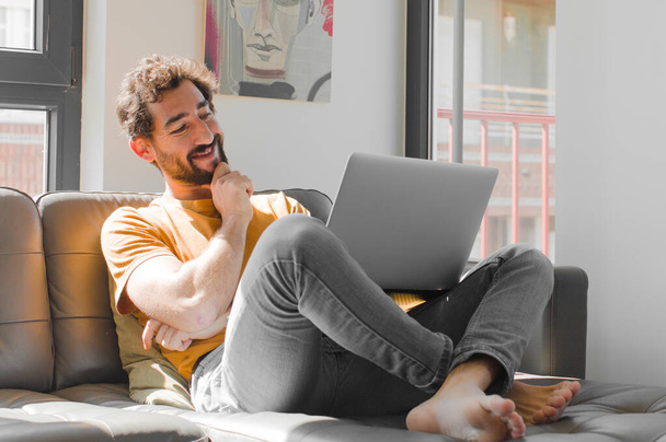 young bearded man smiling with a happy, confident expression with hand on chin, wondering and looking to the side with a laptop on a couch laptop concept - Φωτογραφία, εικόνα