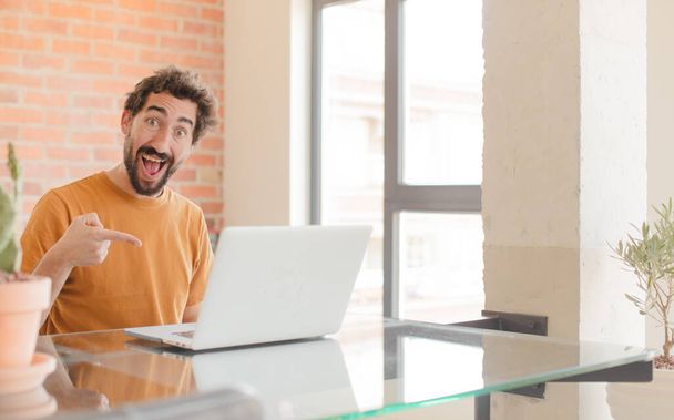 young bearded man looking excited and surprised pointing to the side and upwards to copy space and working with a laptop - Foto, imagen