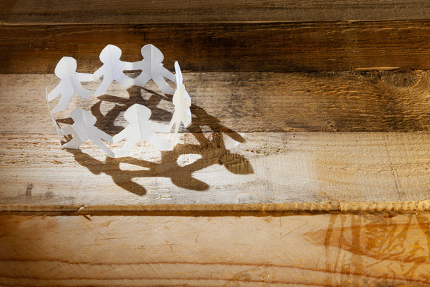 Cercle de figures humaines en papier blanc sur une surface en bois. Une lumière latérale projette une ombre. - Photo, image