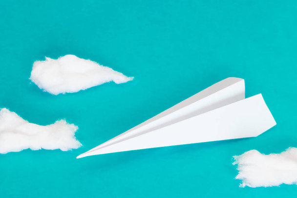 Weißes Papierflugzeug neben einigen Baumwollwolken auf türkisblauem Hintergrund. - Foto, Bild