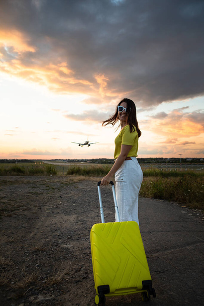 молодая улыбающаяся красивая женщина с желтым багажом посадки самолета на заднем плане. время захода солнца - Фото, изображение