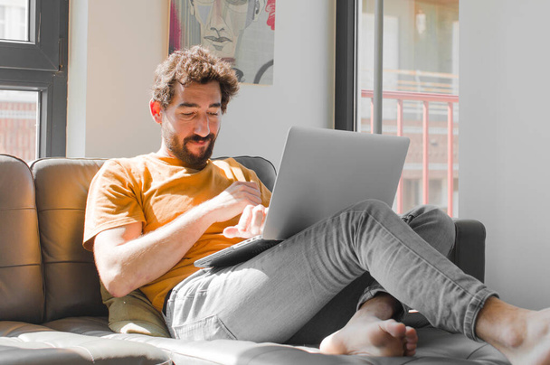 young bearded man shrugging, feeling confused and uncertain, doubting with arms crossed and puzzled look with a laptop on a couch laptop concept - Foto, Imagem