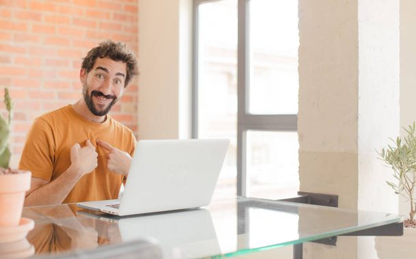 young bearded man feeling happy, surprised and proud, pointing to self with an excited, amazed look and working with a laptop - Foto, immagini