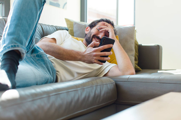 молодой бородатый мужчина отдыхает на диване со своим смартфоном - Фото, изображение