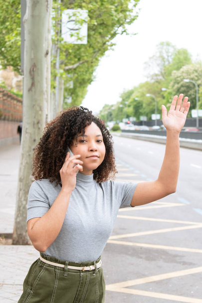 Young black curly haired girl raises her hand to call an uber while talking on her cell phone in the city. Vertical photograph. - Foto, afbeelding