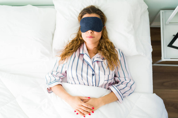 Uykulu kadın beyaz yatağında uzanıyor ve sabahları ışıktan kaçmak için uyku maskesi takıyor. Pijamalı güzel bir kadın yatak odasında uyuyor. - Fotoğraf, Görsel