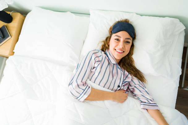 Mujer sonriente despertando por la mañana con una máscara para dormir. Mujer latina feliz sintiéndose bien descansado y acostado en una cama cómoda - Foto, imagen