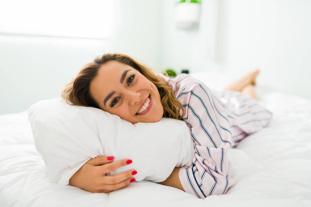 Portrait d'une magnifique femme latine en pjs couchée et reposant sur son lit confortable. Femme souriante étreignant un oreiller blanc et se sentant heureuse - Photo, image