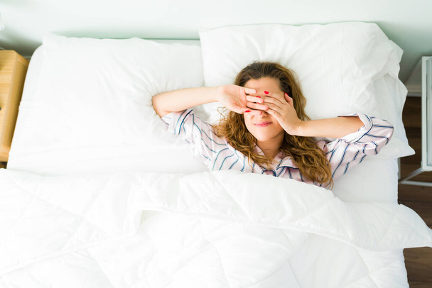 Κουρασμένη ισπανόφωνη γυναίκα ξυπνά στο κρεβάτι της το πρωί και καλύπτει τα μάτια της με τα χέρια της λόγω του έντονου φωτός - Φωτογραφία, εικόνα
