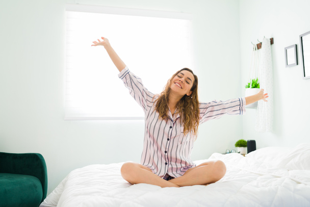 Attractive positive woman with pajamas sitting in bed after waking up and stretching her arms. Hispanic woman feeling happy and well rested - Photo, Image
