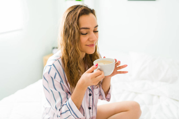 Rahat genç bir kadın pijama giyiyor ve sabah bir fincan kahve içerken yatakta oturuyor. - Fotoğraf, Görsel