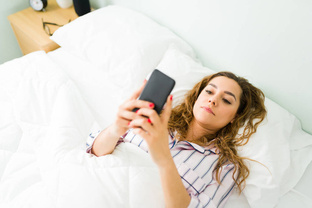 Stanco giovane donna che riposa a letto dopo essersi svegliata la mattina e sms nel suo smartphone - Foto, immagini