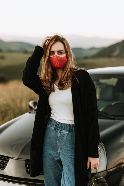 young woman wearing red face mask standing near car in mountains - Fotoğraf, Görsel