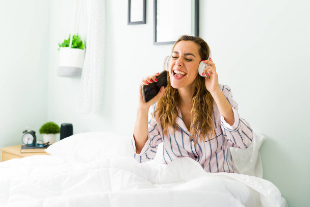 Happy woman singing her favorite song while listening to music with headphones and holding her smartphone. Hispanic woman in pjs having fun in bed - 写真・画像