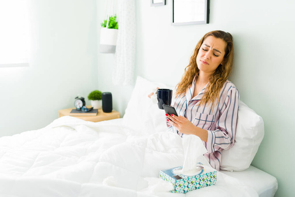 Mulher doente mensagens de texto em seu smartphone enquanto bebe uma xícara de chá quente para sua doença de gripe. Mulher bonita de pijama descansando na cama ao lado de uma caixa de tecidos - Foto, Imagem