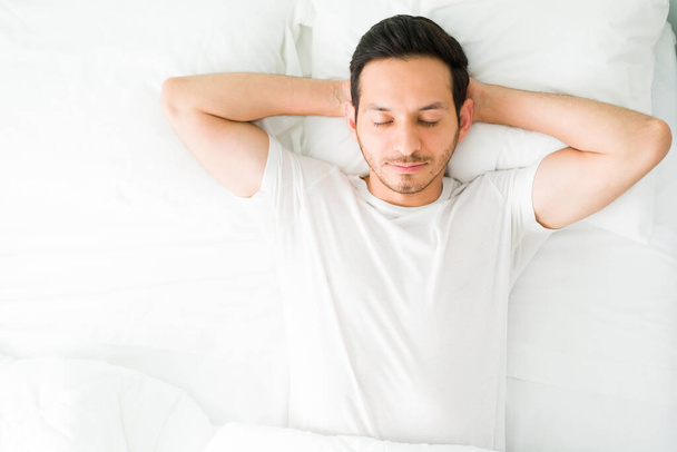 Een knappe Spaanse man rustend op haar schone witte bed met zijn handen achter haar hoofd. Ontspannen man ligt in bed en probeert in slaap te vallen - Foto, afbeelding