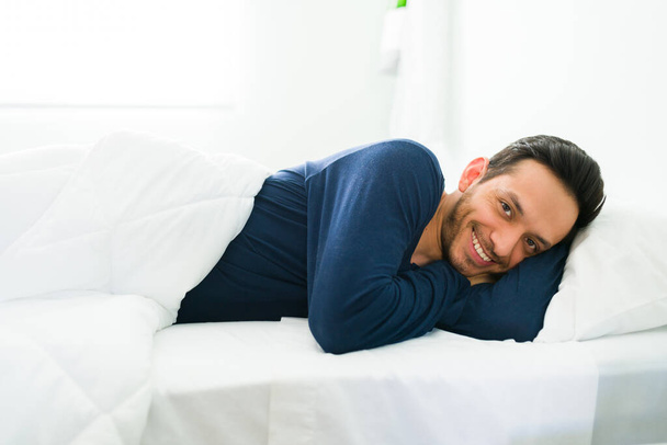 Przystojny mężczyzna nawiązujący kontakt wzrokowy leżąc na wygodnym łóżku i pokryty białą kołdrą. Latynos budzi się rano i uśmiecha - Zdjęcie, obraz