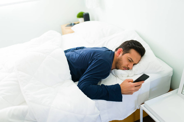 Hispanic man in his 30s lying in bed after waking up while checking his texts and work emails on the smartphone - Foto, imagen