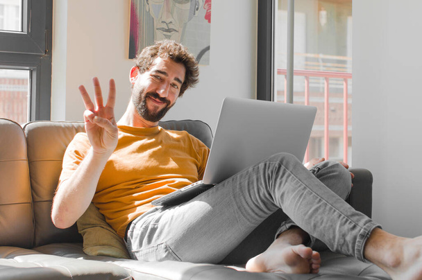 young bearded man smiling and looking friendly, showing number three or third with hand forward, counting down with a laptop on a couch laptop concept - Fotó, kép