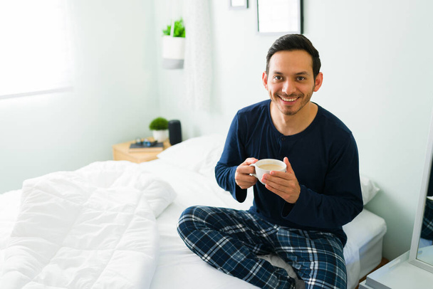 Πορτρέτο ενός όμορφου άνδρα που κάνει οπτική επαφή και χαμογελά πίνοντας ένα φλιτζάνι ζεστό καφέ. Λατίνος με πιτζάμες χαλαρώνει στο κρεβάτι το πρωί - Φωτογραφία, εικόνα