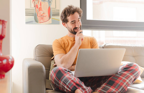 young bearded man smiling happily and daydreaming or doubting, looking to the side and sitting with a laptop - Foto, afbeelding