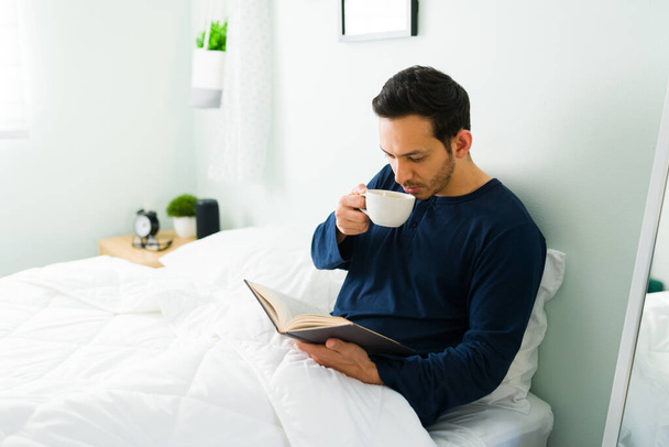 Homem latino atraente desfrutando de sua manhã com uma xícara de café no quarto. Homem inteligente em pjs lendo um livro enquanto descansa em uma cama confortável - Foto, Imagem