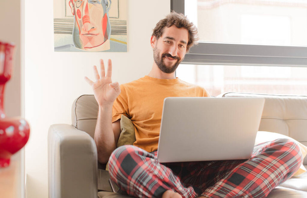 young bearded man smiling and looking friendly, showing number four or fourth with hand forward, counting down and sitting with a laptop - Fotoğraf, Görsel