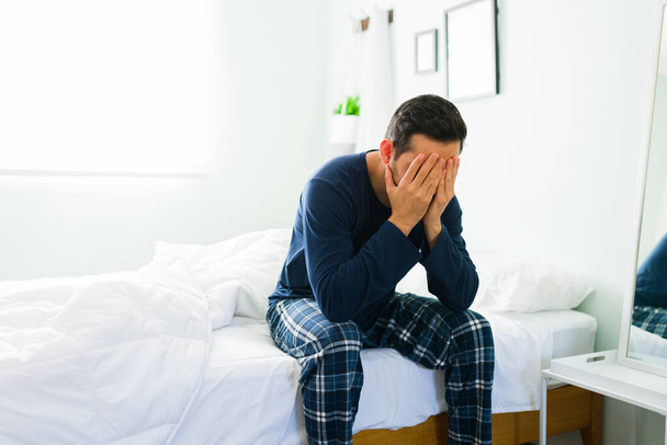 Молодой человек с разбитым сердцем в пижаме, закрывая лицо руками, проснувшись утром. Мужчина чувствует грусть и депрессию в своей спальне - Фото, изображение